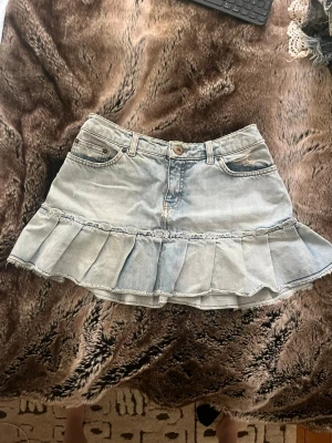 Ljus jeanskjol med volangkant - Säljer en ljusblå jeanskjol med volangkant och klassiska fem fickor. Kjolen har en snygg slitning och knappstängning framtill. Perfekt för dig som gillar en trendig och avslappnad stil.