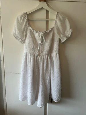 Vit kort klänning med puffärmar - Söt vit kortklänning med puffiga kortärmar och smockad överdel. Klänningen har knytning framtill vid urringningen och är tillverkad i ett lätt, prickigt tyg som ger en romantisk känsla. Perfekt för varma dagar och somriga tillfällen. Klänningens kjol är som en byxa