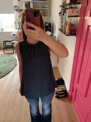 Mörkblå ärmlös randig linnetopp - Säljer en mörkblå ärmlös linnetopp med tunna vita ränder och små röda nyckelpigor. Toppen har en lös passform och rundad halsringning med lätt rynk. Perfekt till jeans för en chill look.