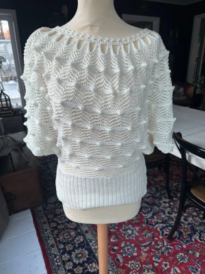 Vit virkad stickad tröja - Säljer en vit stickad tröja med virkat mönster och bred ribbad mudd i midjan. Tröjan har korta, vida ärmar och en unik öppen detalj vid halsringningen. Perfekt för dig som gillar retrostil och luftiga plagg.