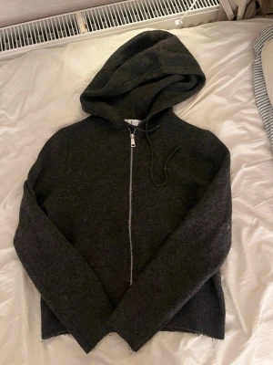 Mörkgrå hoodie med dragkedja - Mysig mörkgrå hoodie med dragkedja framtill och justerbar huva. Tillverkad i ett mjukt, stickat material som känns skönt mot huden. Långa ärmar och ribbade muddar ger en avslappnad look. Perfekt för chill dagar eller när du vill ha något bekvämt.