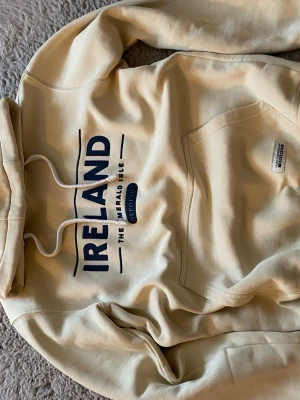 Beige hoodie från Irish Connexxion - Snygg beige hoodie med trycket 'IRELAND THE EMERALD ISLE' i mörkblått framtill. Klassisk känguruficka och justerbar huva med vita snören. Tillverkad i mjukt bomullsmaterial, perfekt för chill dagar. Märkt 'Irish Connexxion Originals' på fickan.