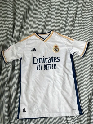 Real Madrid matchtröja Adidas vit - Säljer en vit Real Madrid matchtröja från Adidas med klubbmärke på bröstet och 'Emirates Fly Better' tryckt framtill. Tröjan har guldfärgade detaljer på axlarna, mörkblå ränder på sidorna och är tillverkad i ett lätt, ventilerande material. Perfekt för fotbollsträning eller match.
