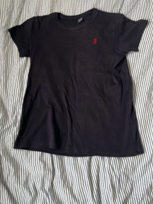 Svart t-shirt från Ralph Lauren - Klassisk svart t-shirt från Ralph Lauren med röd broderad logga på bröstet. T-shirten har rund halsringning och korta ärmar. Perfekt basplagg med stilren design och skön passform i mjuk bomull.