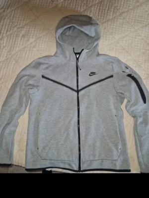 Grå Nike Tech Fleece hoodie - Snygg grå Nike Tech Fleece hoodie med dragkedja och svarta detaljer. Hoodien har huva, två sidofickor med dragkedja och en extra ficka på ärmen. Tillverkad i mjukt och bekvämt material, perfekt för en sportig och avslappnad stil.