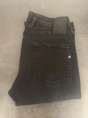 Svarta Replay anbass jeans, stl 33 - Säljer ett par svarta slim fit jeans från Replay med klassisk femficksmodell och diskret logga på bakfickan. Jeansen har normal midja och är tillverkade i stretchigt denimtyg för extra komfort. Perfekta till en avslappnad och stilren look.