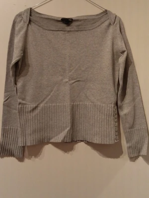 Grå stickad tröja med ribbade detaljer - Grå stickad tröja från H&M med bred ribbad kant nertill och vid ärmslut. Tröjan har rund halsringning och dekorativa knappar på ena sidan. Perfekt för lager-på-lager och enkel att matcha med jeans eller kjol.