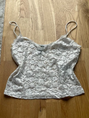 Vit spetslinnetopp med smala band - Vitt spets linne med tunna axelband och blommig spets över hela toppen. Croppad modell.