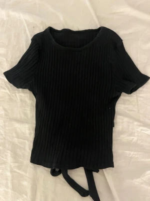 Svart ribbad topp med knytband - Säljer en svart ribbad topp med korta ärmar och rund hals. Toppen har snygga knytband i ryggen som ger en trendig look. Materialet är stretchigt och följsamt, perfekt för att styla till jeans eller kjol.