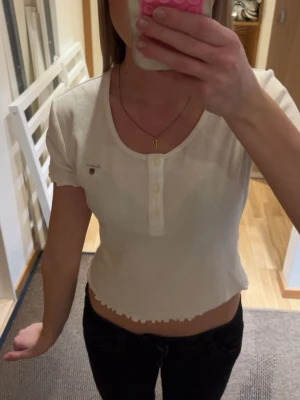 Vit croppad topp från Gant - Säljer en vit croppad topp från Gant med korta ärmar och knappdetaljer framtill. Toppen har en ribbad struktur och vågig kant nertill för en trendig look. Perfekt att matcha med höga jeans eller kjol.