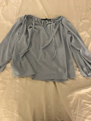 Ljusblå offshoulder blus med band - Trendig ljusblå offshoulder blus med långa ärmar och dekorativa knytband vid halsringningen. Blusen har en lös passform och är tillverkad i ett mjukt, ribbat tyg som ger en avslappnad men stilren look.