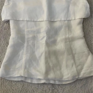 Vit axelbandslös linnetopp - Säljer en stilren vit axelbandslös topp i linnematerial. Toppen har en bandeau-liknande överdel och en figurnära passform. Perfekt att matcha med jeans eller kjol. 