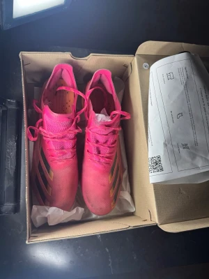 Adidas rosa fotbollsskor med svarta ränder - Säljer ett par snygga fotbollsskor från Adidas i stark rosa färg med svarta och gula detaljer på sidorna. Skorna har snörning och en låg profil för bra bollkontroll. Ovandelen är i syntetmaterial som ger lätt känsla och bra grepp på planen.