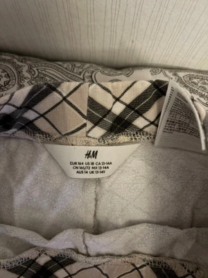Rutiga pyjamasbyxor från H&M - Säljer ett par beige och svart-rutiga pyjamasbyxor från H&M i storlek 164. Byxorna har dragsko i midjan och är tillverkade i mjukt, bekvämt material. Perfekta för mysiga hemmakvällar eller lata morgnar.