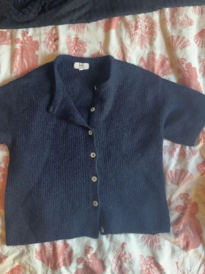 Mörkblå stickad kofta med knappar - Mysig mörkblå stickad kofta med korta ärmar och träknappar framtill. Koftan har en enkel, rak passform och mjukt material som känns skönt mot huden. Perfekt att slänga över en t-shirt eller linne för en avslappnad look.