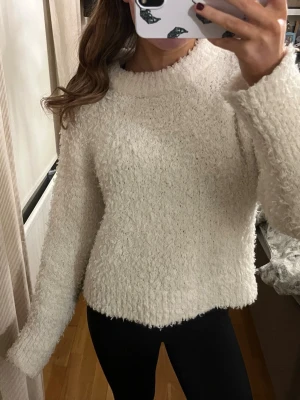 Krämvit fluffig stickad tröja - Mysig krämvit stickad tröja med fluffig textur och hög ribbad hals. Tröjan har långa ärmar och är tillverkad i ett mjukt, luddigt material som känns riktigt skönt mot huden. Perfekt för kyliga dagar när du vill vara både varm och snygg.