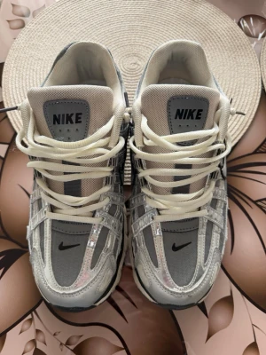 Nike silvergrå sneakers med mesh och skinn - Snygga Nike sneakers i silvergrått med detaljer i mesh och skinn. Skorna har vita snören, svart swoosh på sidan och en chunky sula. Tungan är vadderad och märkt med Nike-logga. Perfekta för dig som gillar sportig och modern stil.