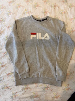Grå sweatshirt från FILA - Snygg grå sweatshirt från FILA med klassisk logga i vitt och rött framtill. Tröjan har rund halsringning, ribbade muddar vid ärmslut och nederkant samt en avslappnad passform. Perfekt för en sportig och casual stil.