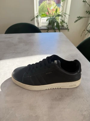 Svarta Axel Arigato sneakers - Stilrena svarta sneakers från Axel Arigato med lågt skaft och vit sula. Skorna är i läder och har diskret logga på sidan. Klassisk siluett med svarta skosnören och minimalistisk design. Perfekta för dig som gillar en clean look. Skorna har några defekter men alla syns på bilderna ovan men inget som syns med ögat om man inte kollar extra noga.