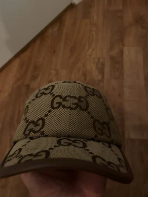 Beige GG keps  - Snygg beige keps med det klassiska GG-mönstret i brunt över hela kepsen. Justerbar rem bak i brunt läder och mesh-liknande material på ovansidan. Perfekt för dig som vill ha en stilren och lyxig accessoar.