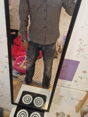 Grå rutig skjorta med knappar - Snygg grå skjorta med diskret rutigt mönster och svarta knappar. Skjortan har lång ärm och klassisk krage, perfekt för en stilren look. Passformen är normal och materialet känns som bomull eller polyester.