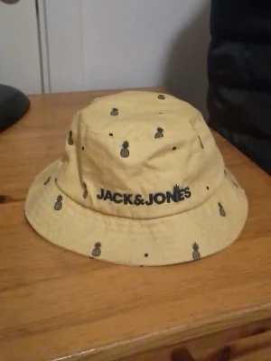 Gul bucket hat med ananas Jack & Jones - Snygg gul bucket hat från Jack & Jones med små svarta ananastryck över hela hatten. Klassisk form med bred brätte och broderad logga framtill. Tillverkad i mjukt tyg, perfekt för soliga dagar och streetstyle.