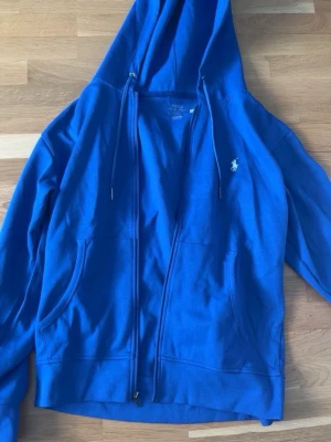 Blå hoodie från Polo Ralph Lauren - Blå hoodie med dragkedja från Polo Ralph Lauren. Klassisk design med huva och dragsnören, två fickor framtill och den ikoniska vita logon broderad på bröstet. Tillverkad i mjuk bomull för skön känsla och avslappnad stil.