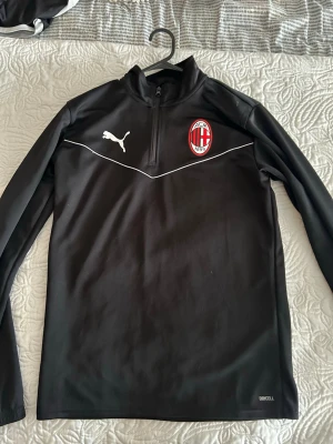 AC Milan träningströja från Puma - Svart långärmad träningströja från Puma med AC Milan-logga på bröstet och vit detalj över bröstet. Tröjan har halv dragkedja och är tillverkad i funktionsmaterial med DryCell-teknologi.