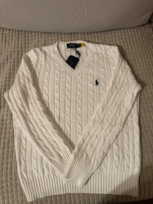 Vit kabelstickad tröja Polo Ralph Lauren - Kabelstickad vit tröja från Polo Ralph Lauren med V-ringning och klassisk marinblå logga broderad på bröstet. Tröjan har långa ärmar och ribbade muddar vid ärmslut och nederkant. Perfekt för en clean och stilren look.