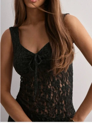 Svart spetslinne med knyt fram - Snyggt svart ärmlöst linne i spets med knyt fram och v-ringning. Toppen är genombruten och har en figurnära passform, perfekt för att skapa en trendig look. Materialet är lätt och stretchigt. 