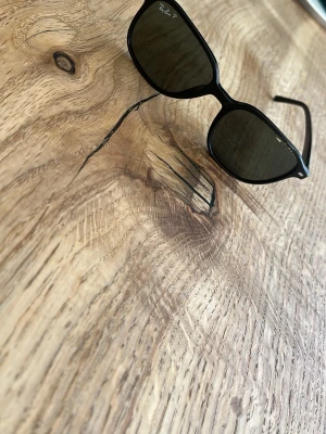 Svarta Ray-Ban solglasögon - Snygga svarta solglasögon från Ray-Ban med klassisk rektangulär form och mörka glas. Bågarna är tunna och stilrena, perfekta för att lyfta din look. Ray-Ban-loggan syns tydligt på sidan av bågen och i hörnet av glaset.