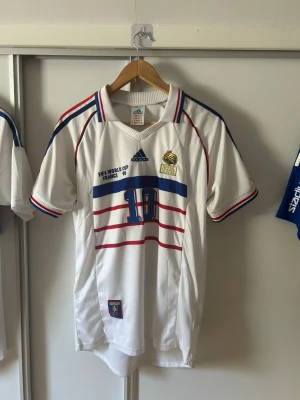 Frankrike Zidane VM 98 matchtröja - Klassisk vit fotbollströja från Adidas, Frankrikes landslag, VM 1998. Nummer 10 och Zidane på ryggen, blå och röda detaljer, broderad tupp-logga och FIFA World Cup-märke. Korta ärmar, rund hals och sportig passform. Perfekt för fotbollsfans!