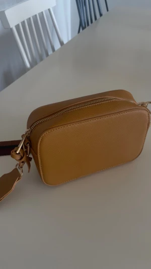 Kamelbrun axelväska med bred rem - Snygg kamelbrun axelväska i slätt konstläder med rektangulär form. Väskan har en bred, justerbar axelrem i vinrött, brunt och svart tyg med guldfärgade metalldetaljer. 