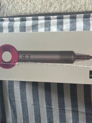 Dyson Supersonic hårfön - Säljer en Dyson Supersonic hårfön i snygg mörkgrå och rosa design. Kommer med flera munstycken för olika stylingbehov. Hårfönen har en modern, rund form och är tillverkad i plast och metall. Perfekt för dig som vill ha snabb och effektiv hårtorkning.