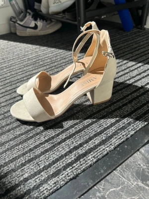 Beige klackpumps från Amisu - Snygga beige pumps från Amisu med öppen tå, bred klack och justerbar ankelrem. Skorna har en stilren design i mockaimitation och passar perfekt till både jeans och klänning. Klacken är stabil och ger en trendig look.