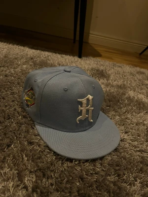 Ljusblå snapback keps med broderad R - Snygg ljusblå snapback keps med stor broderad vit R på framsidan och patch på sidan. Klassisk platt skärm och justerbar baksida. Tillverkad i tåligt bomullstyg, perfekt för en streetwear-look.