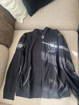 Svart hoodie från SportMaster XL - Svart hoodie från SportMaster i storlek XL med dragkedja framtill och coola tryckdetaljer på bröstet och ärmen. Hoodien har fickor med dragkedja och en sportig vibe, perfekt för chill eller träning.