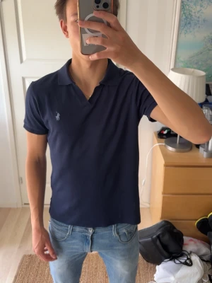 Marinblå pikétröja från Polo Ralph Lauren - Snygg marinblå pikétröja från Polo Ralph Lauren med klassisk krage och korta ärmar. Tröjan har den ikoniska loggan broderad på bröstet och är gjord i mjuk bomull. Perfekt för en stilren och avslappnad look.