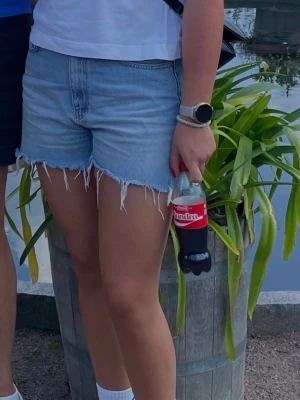 Ljusblå jeansshorts från NA-KD - Säljer ett par ljusblå jeansshorts från NA-KD med råa, fransiga benslut och klassisk femficksdesign. Shortsen har hög midja och är tillverkade i bomull för en skön och avslappnad look. Perfekta för varma dagar och enkel att styla med allt.