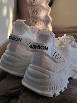 Vita chunky sneakers med meshdetaljer - Snygga vita chunky sneakers med grov sula och stickad mesh på ovandelen. Skorna har vita snören och en plastdetalj med texten 'FASHION' bak på hälen. Perfekta för dig som gillar en sportig och trendig look.