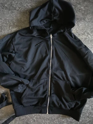 Svart zip-hoodie  - En klassisk svart hoodie med dragkedja framtill och två stora fickor. Hoodien har huva och ribbade muddar vid ärmslut och nederkant. Perfekt för en chill och avslappnad stil. Materialet känns mjukt och bekvämt, troligtvis bomull eller bomullsblandning.