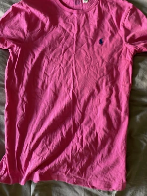 Rosa t-shirt från Polo Ralph Lauren - Snygg rosa t-shirt från Polo Ralph Lauren i modellen Custom Slim Fit. T-shirten har rund halsringning och den klassiska blå broderade loggan på bröstet. Tillverkad i mjuk bomull som känns skön mot huden.