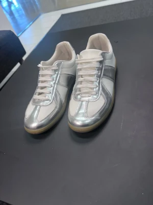 Vita och silverfärgade Maison margiela skor  - Snygga sneakers i vitt och silver med klassisk låg profil och gummisula. Skorna har vita skosnören och paneler i metallic-silver som ger en futuristisk vibe. Perfekta för dig som vill sticka ut med en stilren men ändå unik look.