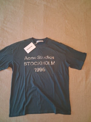 Acne Studios blå t-shirt M - Bla -shirt från Acne Studios med tryck 'Acne Studios STOCKHOLM 1996' på bröstet. Klassisk passform med rund hals och korta ärmar. Tillverkad i mjuk bomull, perfekt för en stilren och avslappnad look.