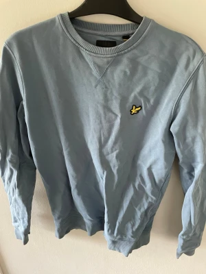 Blå sweatshirt från Lyle & Scott - Ljusblå sweatshirt från Lyle & Scott med klassisk rund halsringning och ribbade muddar. Tröjan har det ikoniska gula fågelmärket broderat på bröstet. Tillverkad i mjuk bomull för en skön och avslappnad look.