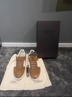 Valentino Garavani Flycrew - Snygga sneakers från Valentino i brunt mocka och vitt läder. Klassisk låg modell med vita sulor och bruna skosnören. Perfekta för dig som vill ha en stilren och exklusiv look. Kommer med originalpåse och box. 13 nitar, mockan fram på sulan är lite smått sliten men går säkert att borsta upp😊