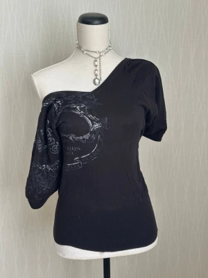 Svart oneshoulder topp med tryck - Cool svart oneshoulder topp med tryck i silver på framsidan. Toppen har en unik asymmetrisk design med en ärm och bred mudd nedtill. Materialet känns mjukt och stretchigt, perfekt för en trendig och edgy look. 
