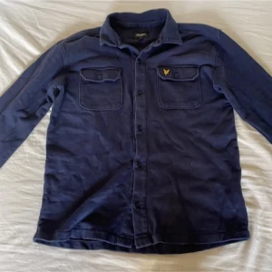 Mörkblå overshirt från Lyle & Scott - Mörkblå overshirt (Skjorta) från Lyle & Scott Junior med klassisk krage, två bröstfickor med lock och knapp samt gul logga på vänster ficka. Skjortan har långa ärmar och knäppning framtill. Perfekt för lager på lager och en stilren look.