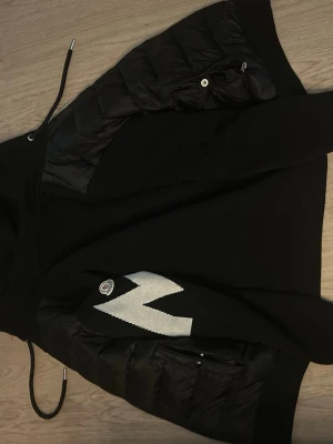 Svart cardiganjacka från Moncler - Snygg svart cardiganjacka från Moncler med quiltade partier och dragkedja. Jackan har en stor vit M-detalj på framsidan och Moncler-logga. Materialmix med stickad ull och glansiga paneler ger en modern look. Perfekt för dig som vill ha något både stilrent och trendigt. 