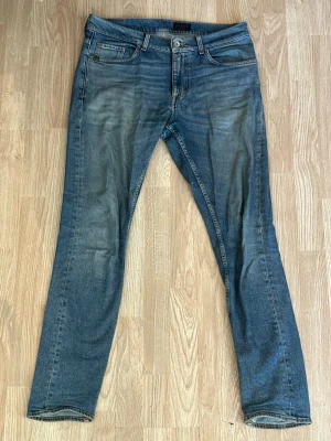Blå slim fit jeans från Tiger of Sweden - Säljer ett par blå slim fit jeans från Tiger of Sweden med klassisk femficksmodell och snygga slitningar framtill. Jeansen har normal midja, dragkedjegylf och patch med logga bak i midjan. Perfekta för en avslappnad och trendig look.